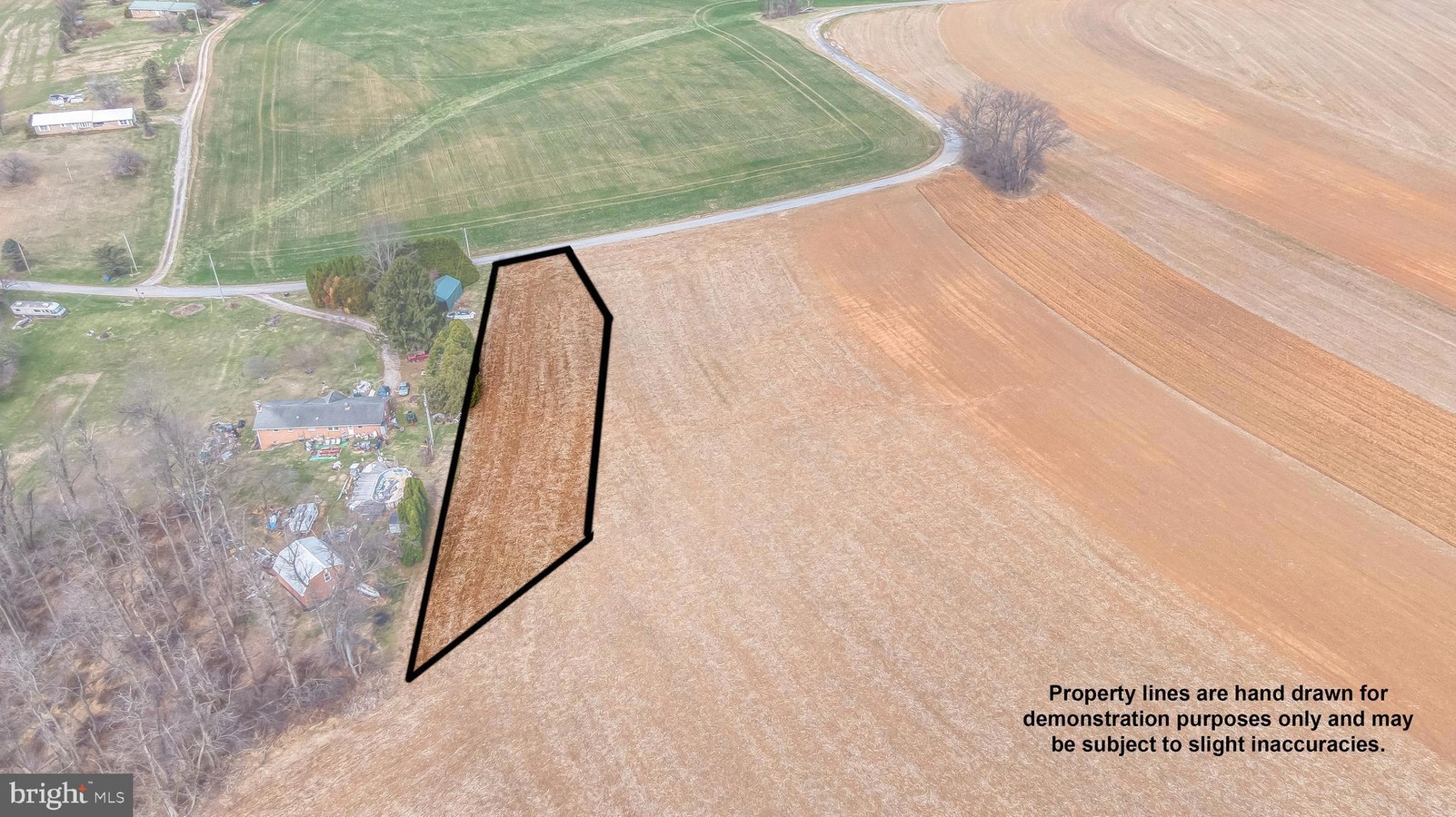 Aerial view of a rural property with an irregular black outline marking the parcel along a field and trees beside a house and road, showing the boundary.