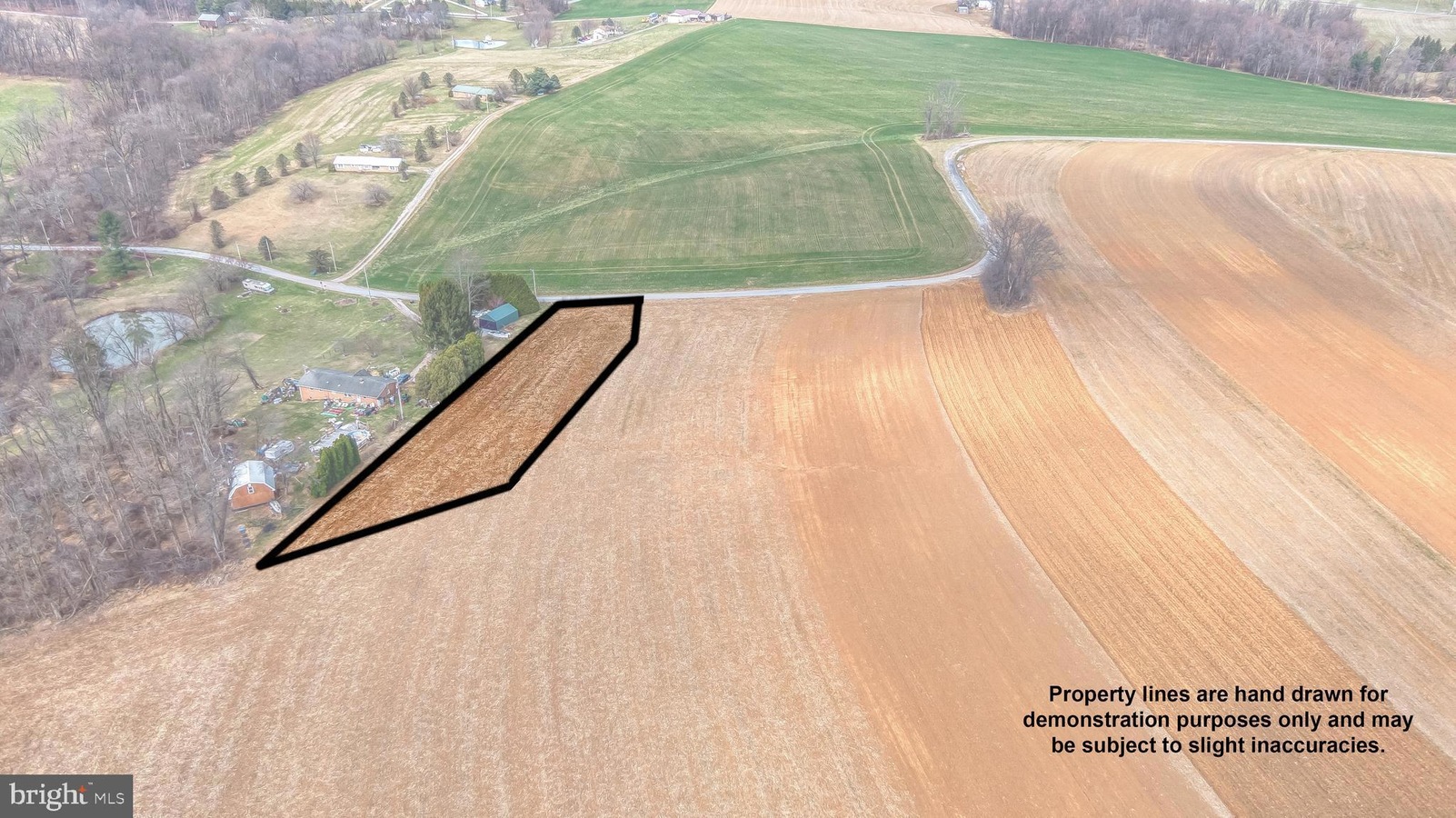 Aerial view of farmland with an irregular black-outlined property boundary along a road, near houses and trees and a small wooded area in the left.