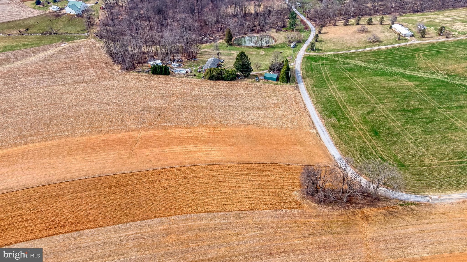 Aerial view of a rural farm with brown harvested fields, a curved road, and a cluster of houses at the treeline.