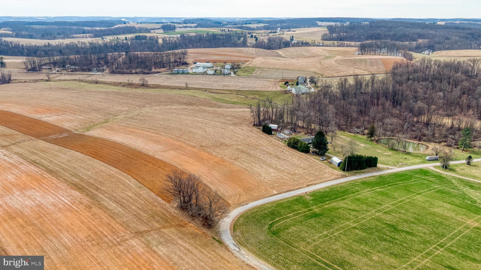Aerial view of a rural farm landscape with patchwork brown and green fields, a curved road, and clusters of houses among leafless trees in early spring.