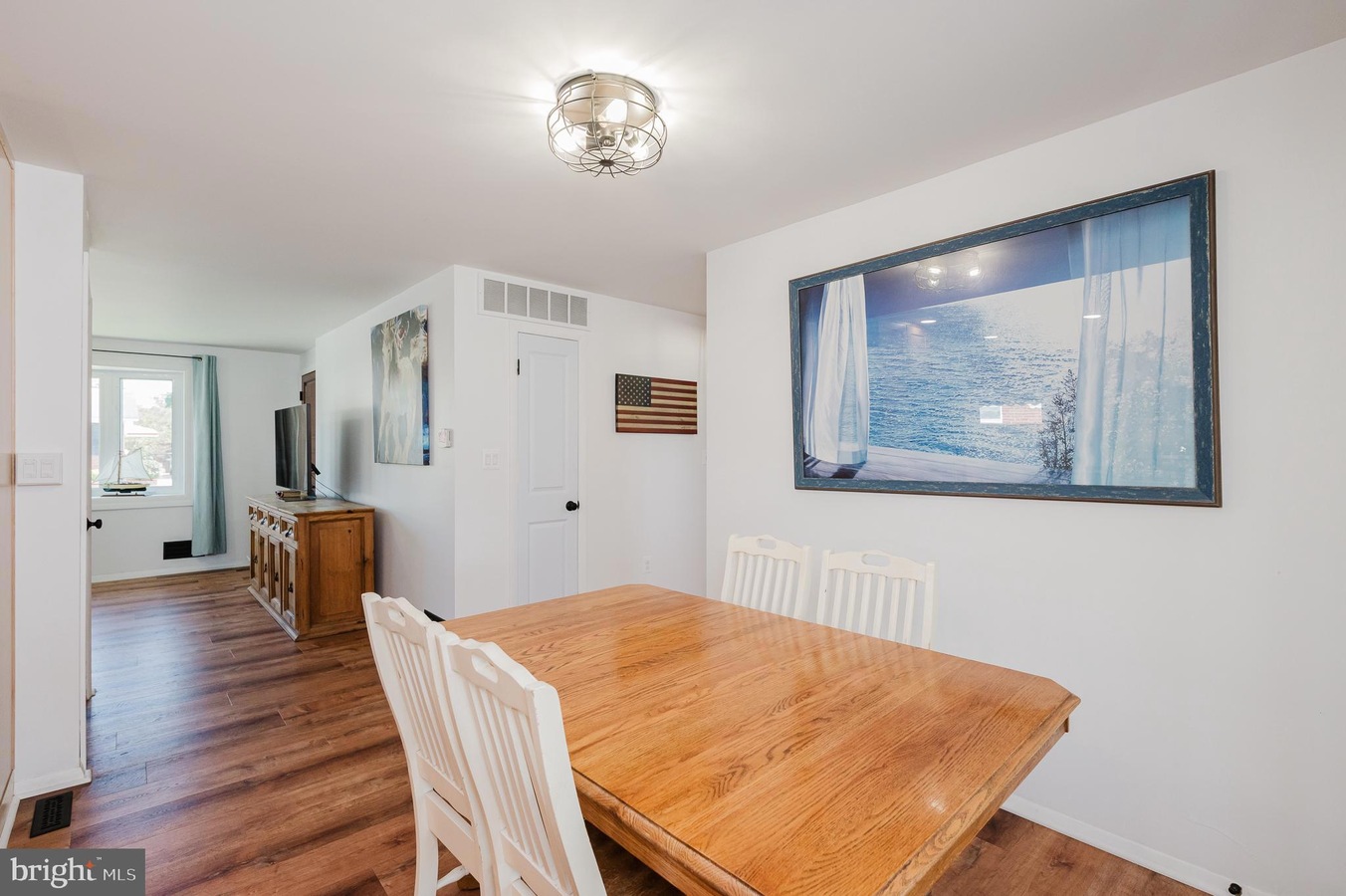 Dining room with a wooden table and white chairs; large ocean-view artwork on the wall and an American flag nearby.