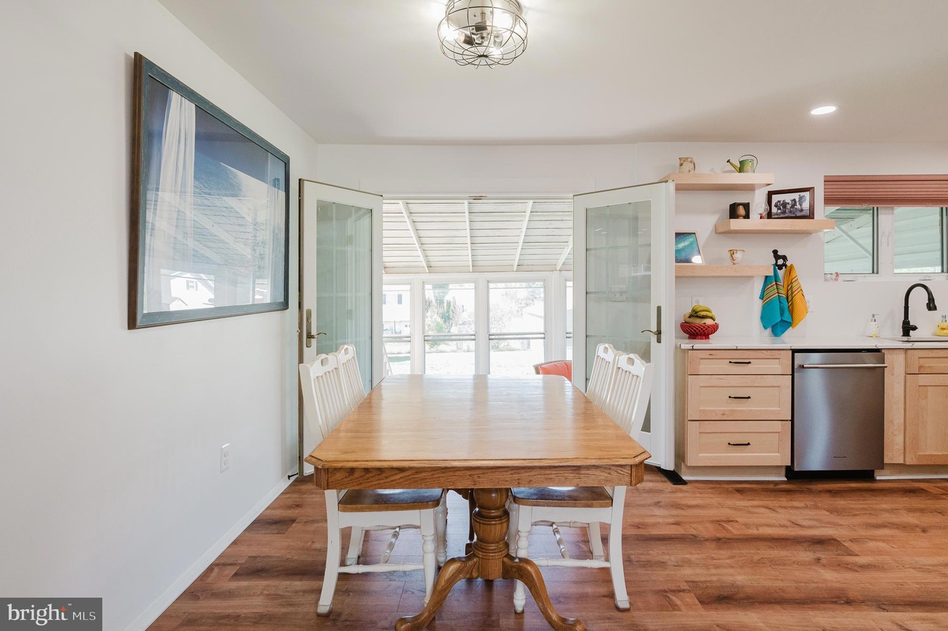 Bright dining area with a wooden table and white chairs, open French doors leading to a sunlit room beyond.