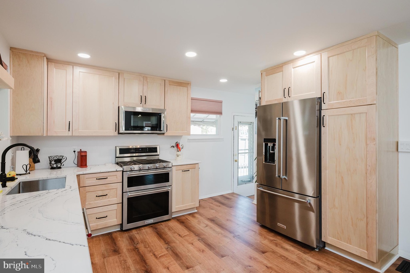 Bright kitchen with light wood cabinets, stainless steel fridge, and double ovens against a white wall. Sturdy hardwood floors run through the space.] ,