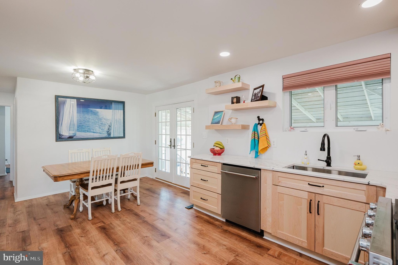 Bright kitchen and dining area with a wooden dining table and white chairs, light wood cabinets, and a stainless dishwasher beneath a marble-like counter.