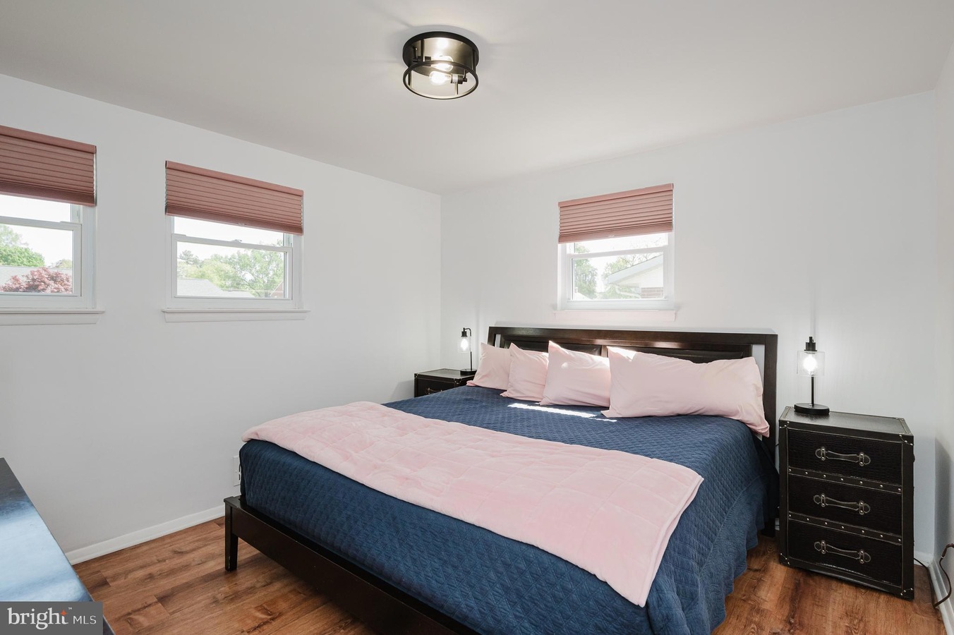Bedroom with a dark wood bed, navy quilt, and pale pink pillows; pink throw at foot and matching blinds in three windows.
