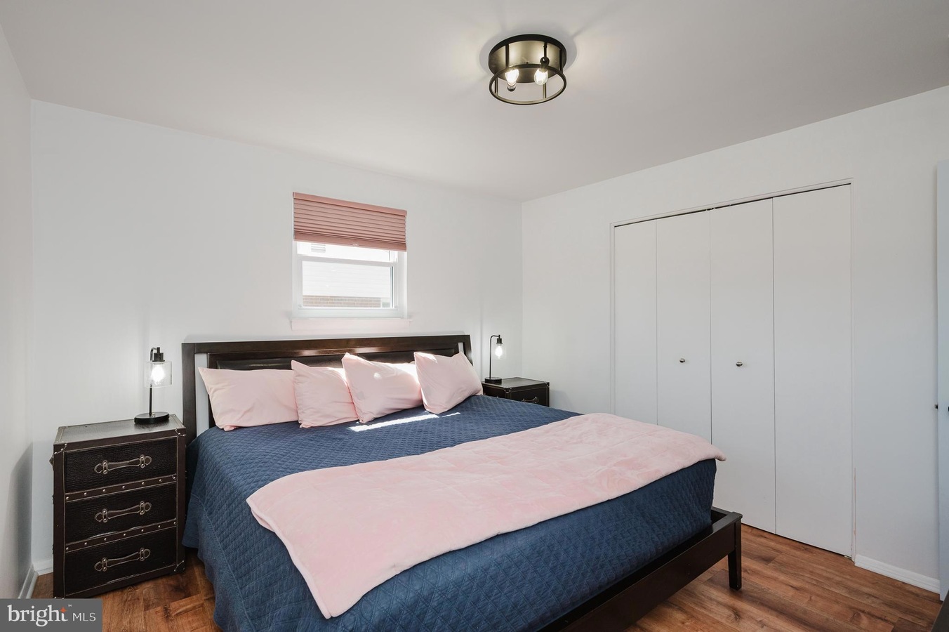 Bedroom with a dark wood bed, navy quilt, pink pillows and throw, two matching nightstands with lamps, small window with a pink shade, and white double closet doors.