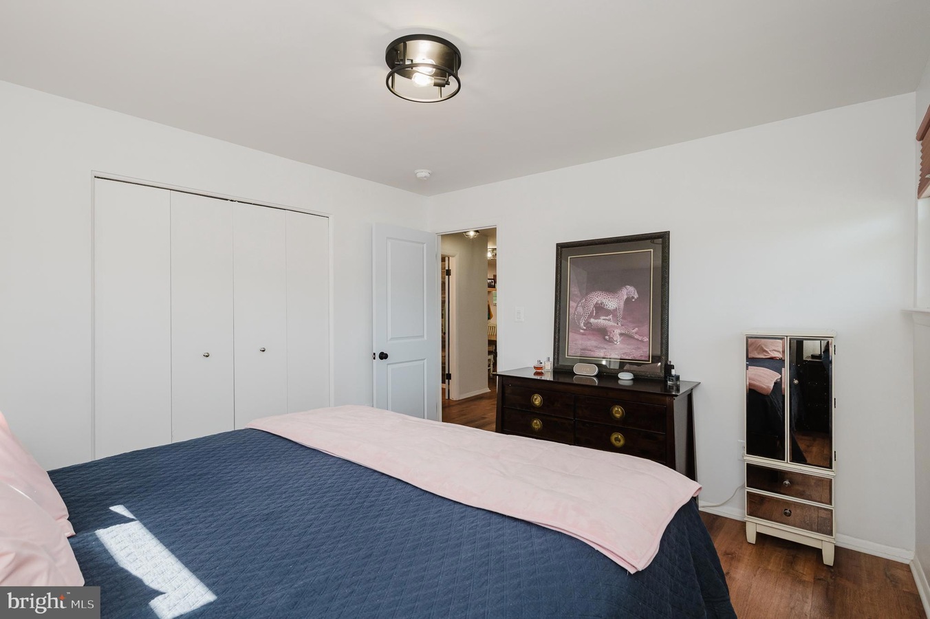 Bedroom with a blue quilt, pink throw, and white walls; a dark wooden dresser sits against the wall with a framed leopard art print above it.