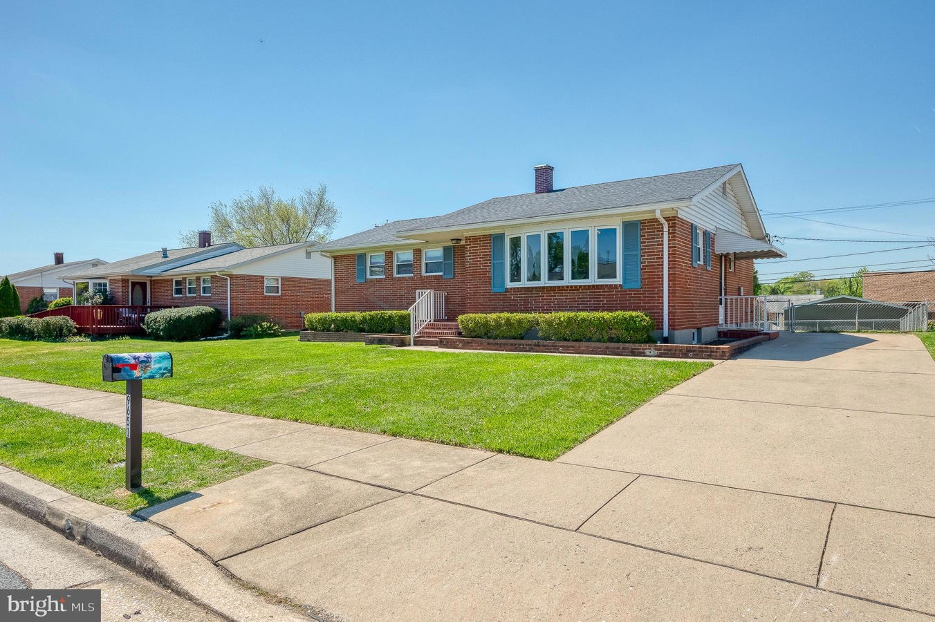 Brick ranch-style house with a manicured front lawn, hedges, and a long driveway on the right under a clear blue sky.