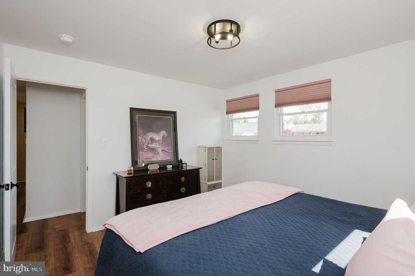 Bedroom with white walls, a dark dresser topped by a framed leopard print, and a bed covered in a navy quilt with a pink throw. Limit: concise and informative for screen readers.