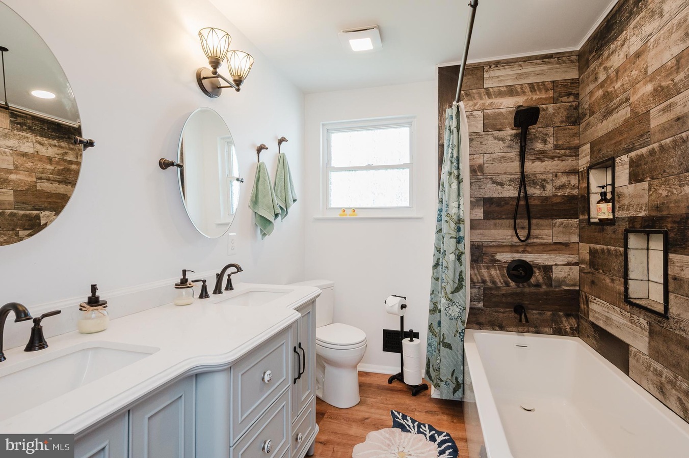 Bright bathroom with a light blue double-sink vanity, two round mirrors, and black fixtures next to a bathtub/shower combo with wood-paneled walls and a blue shower curtain