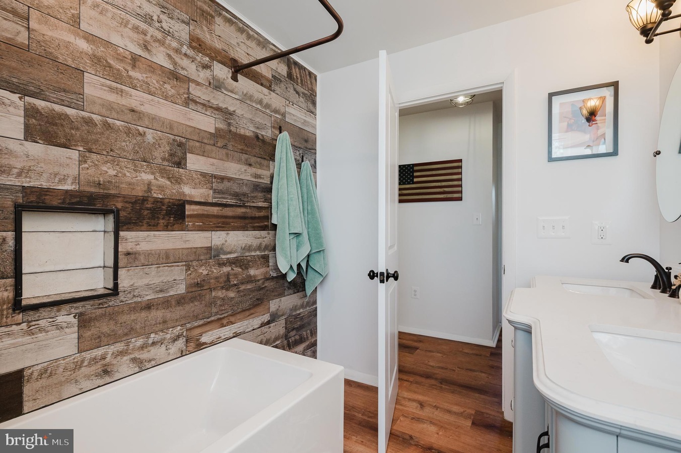 Bathroom with white tub and rustic wood plank wall; teal towels on hooks, and a curved white vanity with a sink nearby.