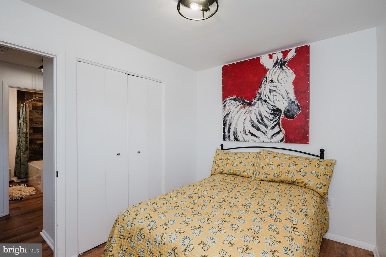 Small bedroom with white walls and a twin bed in a yellow floral quilt, black metal headboard, and a bold red zebra painting above the bed.