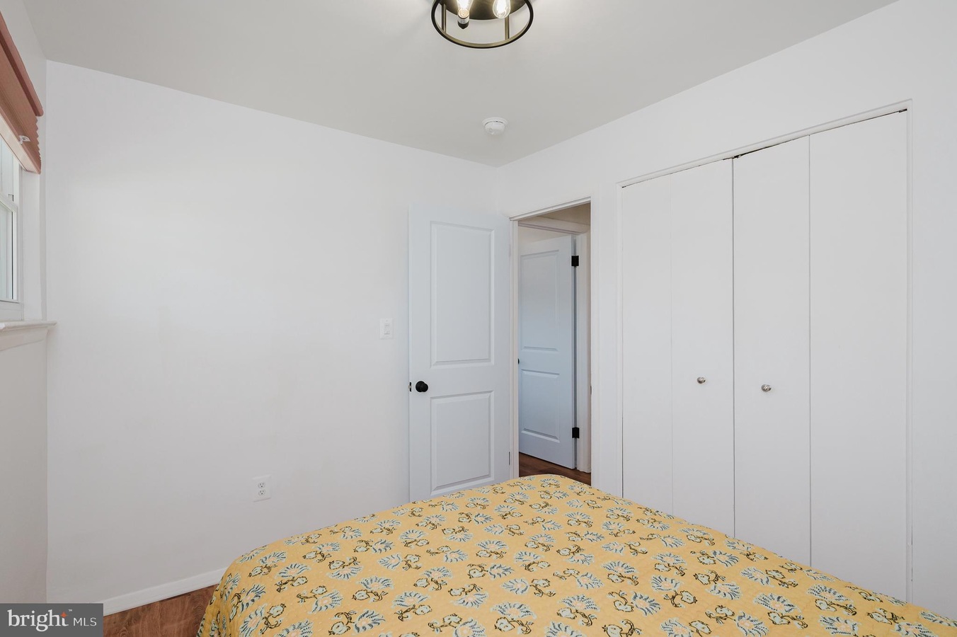 Bright, minimalist bedroom with white walls, a bed in the foreground covered by a yellow floral quilt, and white closet doors along one wall; a doorway leads to another room; small window on the left side.