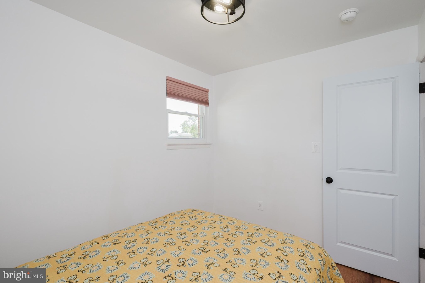 Small white bedroom with a bed covered in a yellow floral quilt, a window with pink blinds, and a white paneled door to the right. A ceiling light hangs above.