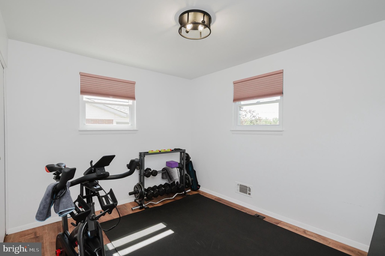 Small home gym in a bright white room featuring a stationary bike, a dumbbell rack with weights, a black exercise mat, and a few yoga accessories by the wall beneath two pink-blind windows.
