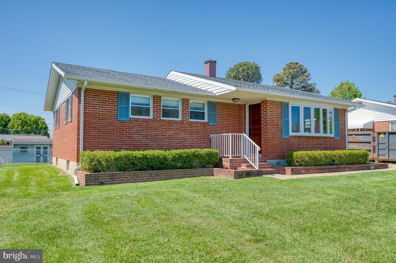 Front view of a single-story brick ranch-style house with blue shutters, white railing, and a brick staircase leading to the front door, surrounded by a green lawn and trimmed hedges.