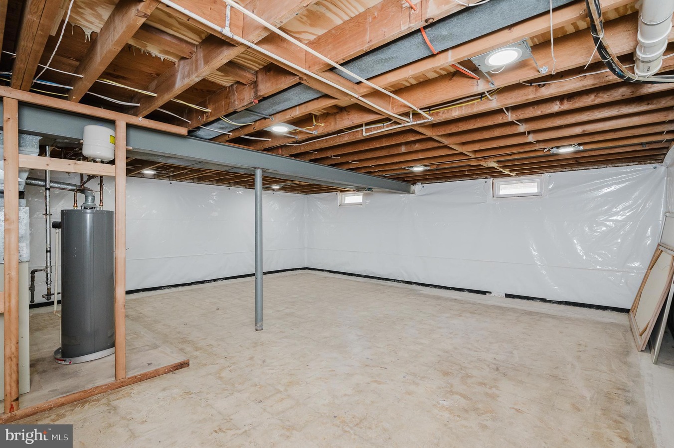 Unfinished basement with exposed joists, a grey water heater, a vertical support post, and white plastic-wrapped walls.