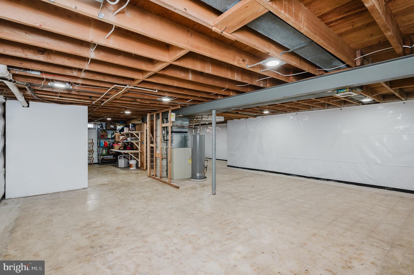 Basement with exposed wooden ceiling beams, a storage area, and a water heater against a white wall. sharp concrete floor.