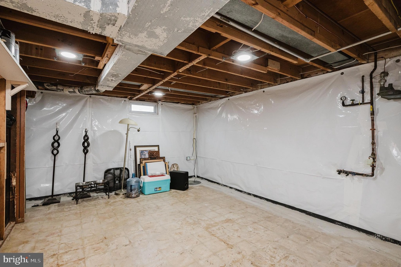 Unfinished basement with plastic-walled sides, exposed ceiling beams, and stored items like a cooler, water jug, lamp, and framed pictures.]','Basement corner with a floor lamp, cooler, water jug, and stacked framed artwork against plastic walls.','Utility basement scene showing exposed pipes on the right and various storage items scattered around.','Cluttered basement storage area with a standing lamp, cooler, water jug, and several frames propped against the wall.','A spacious basement room with white plastic sheeting on the walls, concrete floor, and miscellaneous belongings.