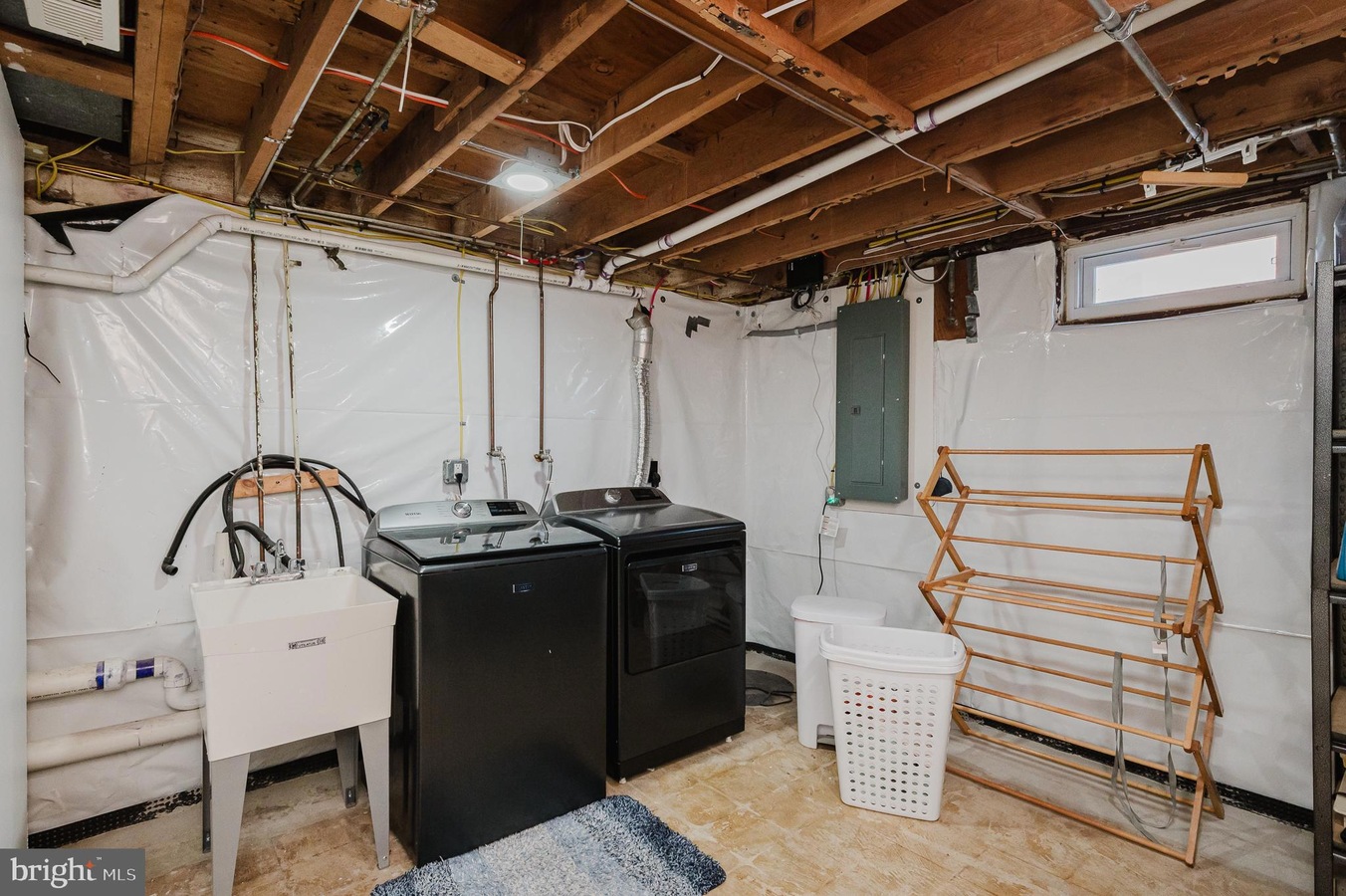Basement laundry area with a washer and dryer, utility sink, laundry basket, and a wooden drying rack against white plastic-wrapped walls.