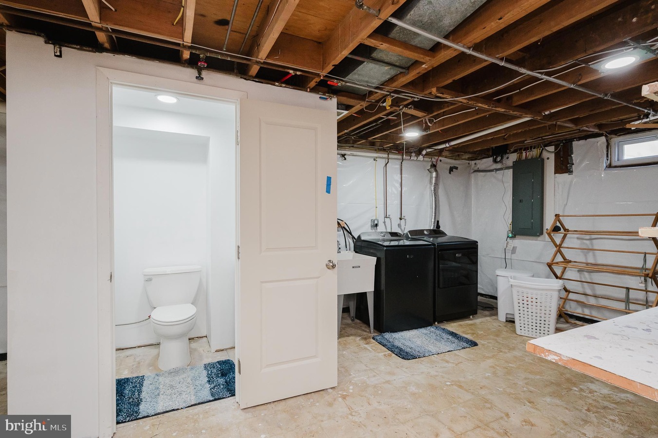 Basement utility area with a washer and dryer, utility sink, laundry basket, exposed ceiling joists, and a small bathroom with a toilet nearby.