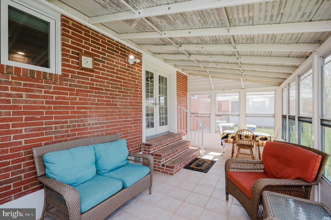 Sunroom with brick wall, teal cushions on a wicker sofa, red-cushioned chair, and a glass-top table by a row of windows.