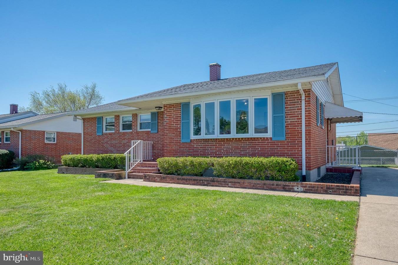 Front view of a brick ranch home with blue shutters, a large bay window, and neatly trimmed hedges along the front. The lawn is green and the walkway leads to brick steps.