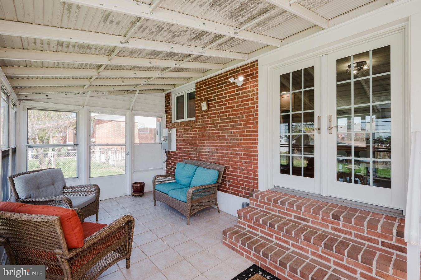 Brick-walled screened porch with wicker seating and turquoise cushions, brick steps to glass French doors on a sunny day behind white frames.