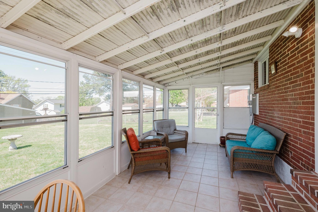 Sunroom or enclosed porch with wicker chairs, turquoise cushions, and a brick wall, facing a grassy backyard through large windows.