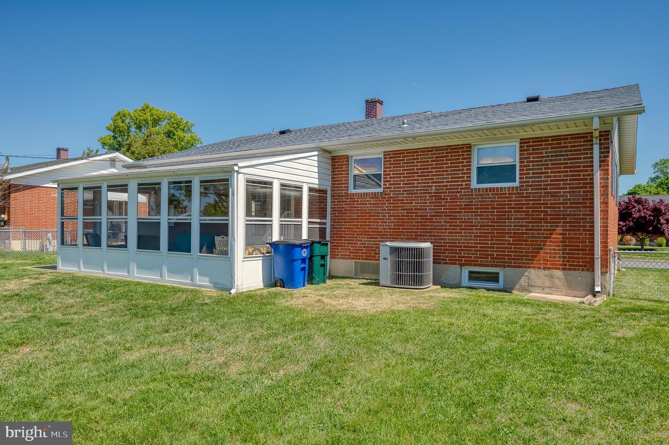 Backyard of a brick house with a white screened porch and blue/green trash bins on the grass in bright daylight.