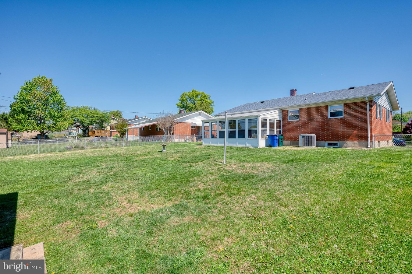 Backyard of a brick house featuring a glass-enclosed sunroom, a green lawn, and a clear blue sky; chain-link fence and trees in the yard.