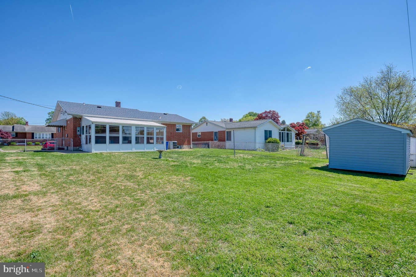 Backyard scene with a brick house featuring a white sunroom, a fenced lawn, and a gray shed on the right under a clear blue sky.