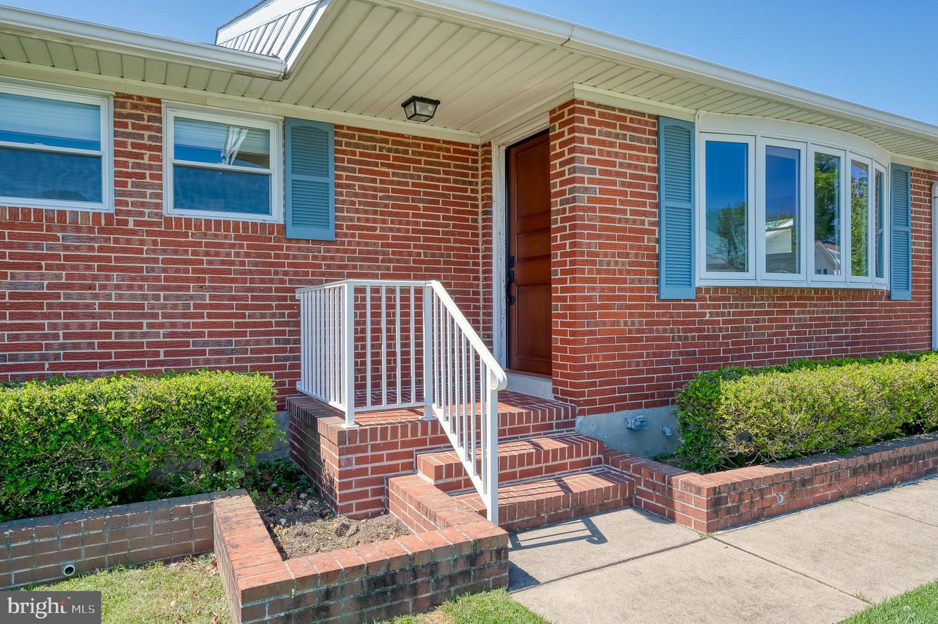 Brick ranch-style home with a white railing and brick steps leading to a wooden front door; blue shutters frame the windows on the brick facade, with trimmed hedges along the walkway.