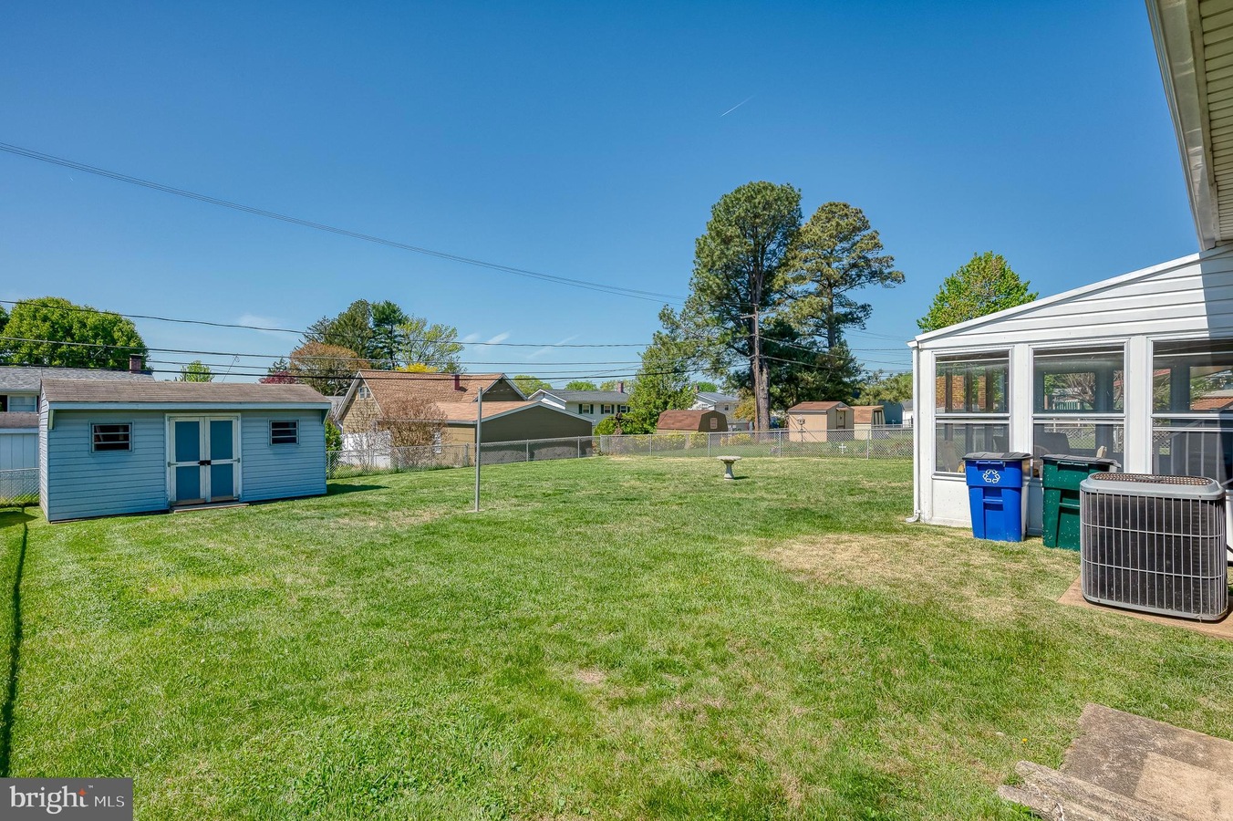 Backyard scene: a small blue shed on the left, a sunroom with several trash bins on the right, a fence, and tall trees in the distance under a clear blue sky.