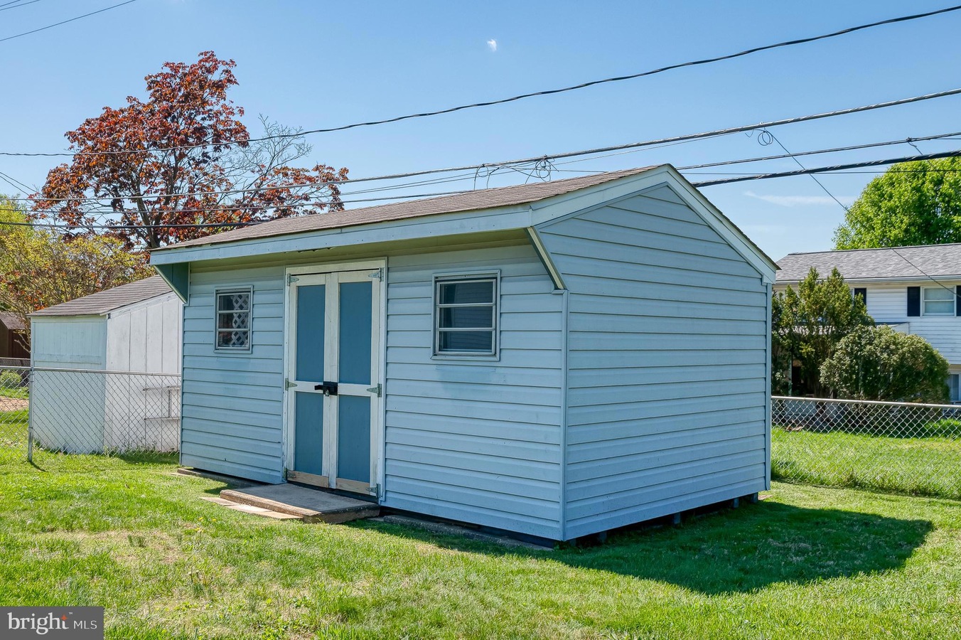 Small blue wooden shed with double doors in a grassy yard, chain-link fence behind, and overhead power lines in a suburban setting.