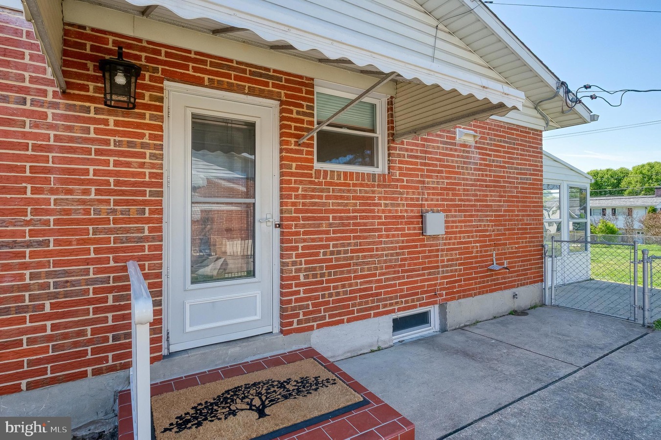 Brick house entry with a white door, small side window, and a tree-pattern doormat on a brick stoop under a white awning.