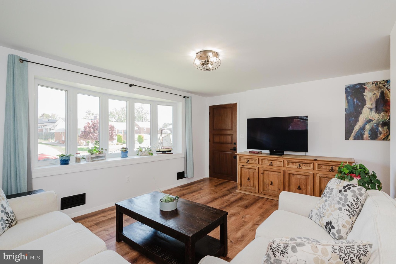 Bright living room with a large bay window, white sofa and spacious wooden coffee table facing a TV on a wooden sideboard.