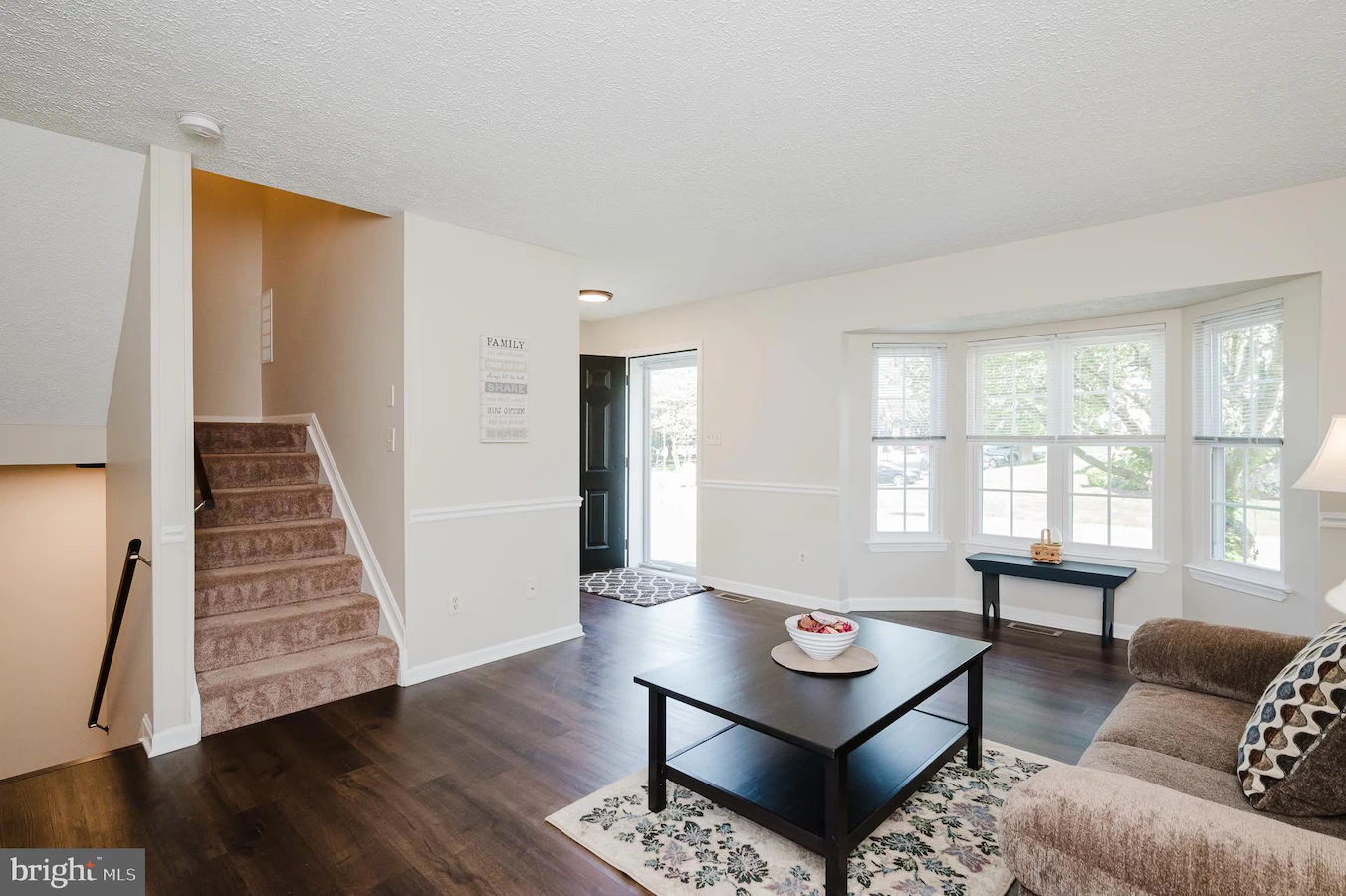 Bright living room with dark wood floors, beige sofa, black coffee table on a patterned rug, and a bay window; stairs lead to the left behind a wall.
