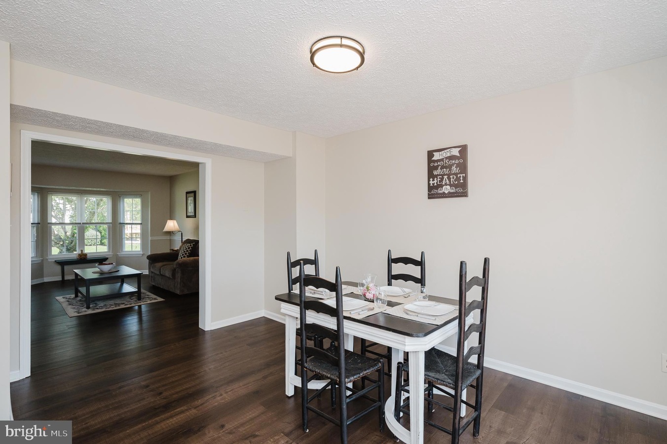 Dining area with a white table set for four and dark chairs, beige walls, and a wall sign reading 'Home is where the heart is' with a living room visible beyond the doorway.