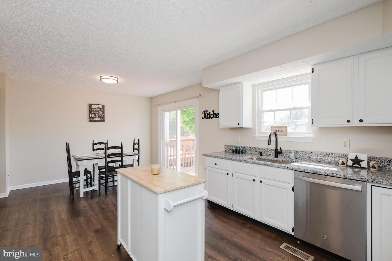 Bright kitchen with white cabinets, granite countertops, island, and a stainless dishwasher, adjacent to a dining table area.