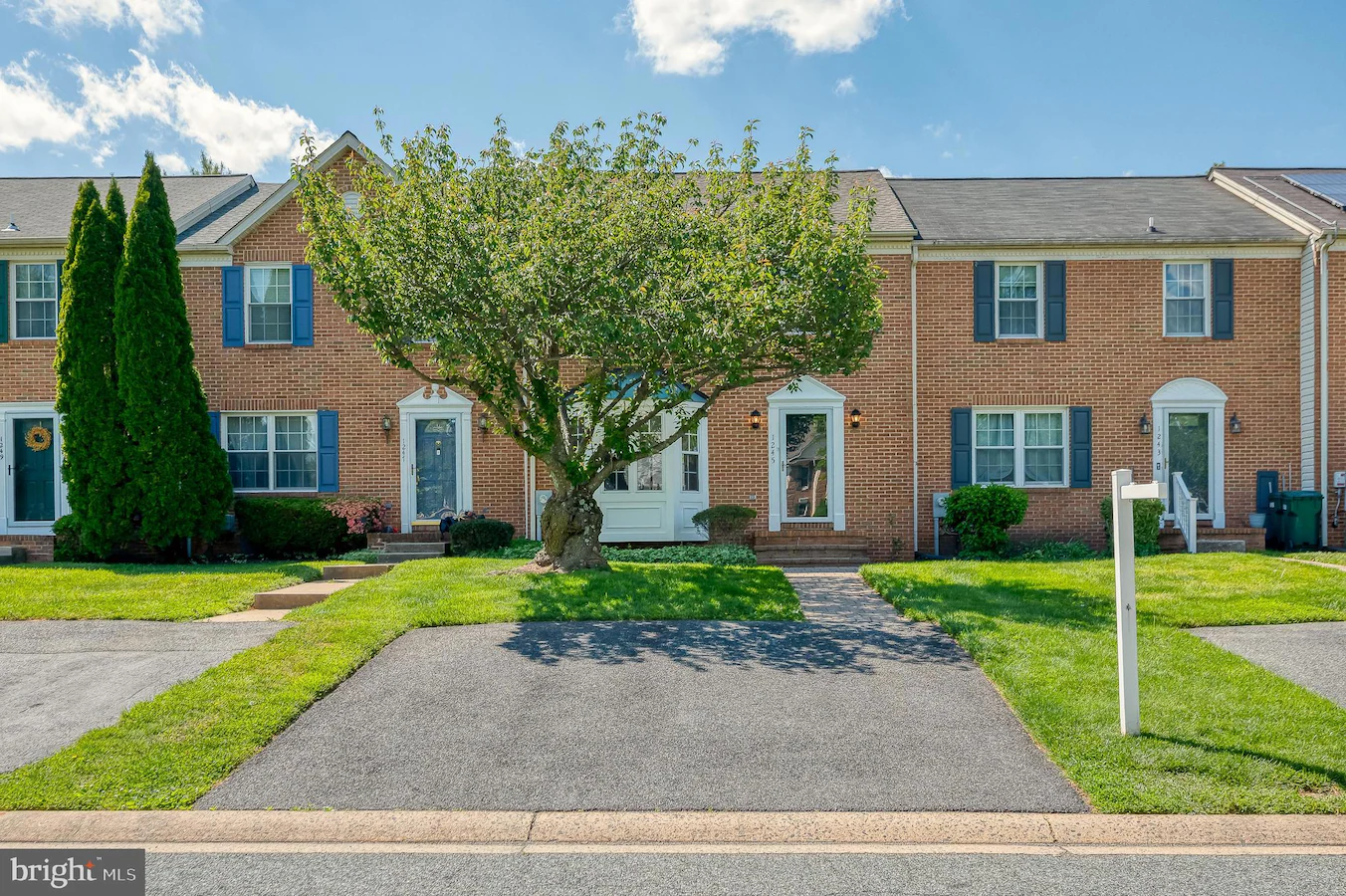 Brick townhouses with blue shutters line a sunny suburban street; a large tree dominates the front yard of the center home, with driveways and well-kept lawns nearby.