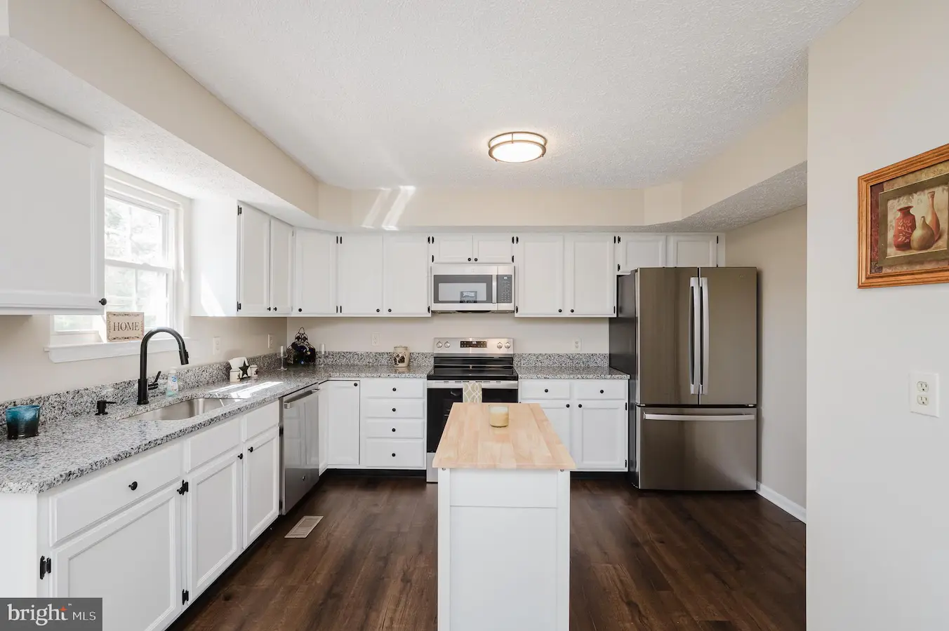 Bright kitchen with white cabinets, granite countertops, and a central island; stainless steel fridge and stainless appliances along the back wall.