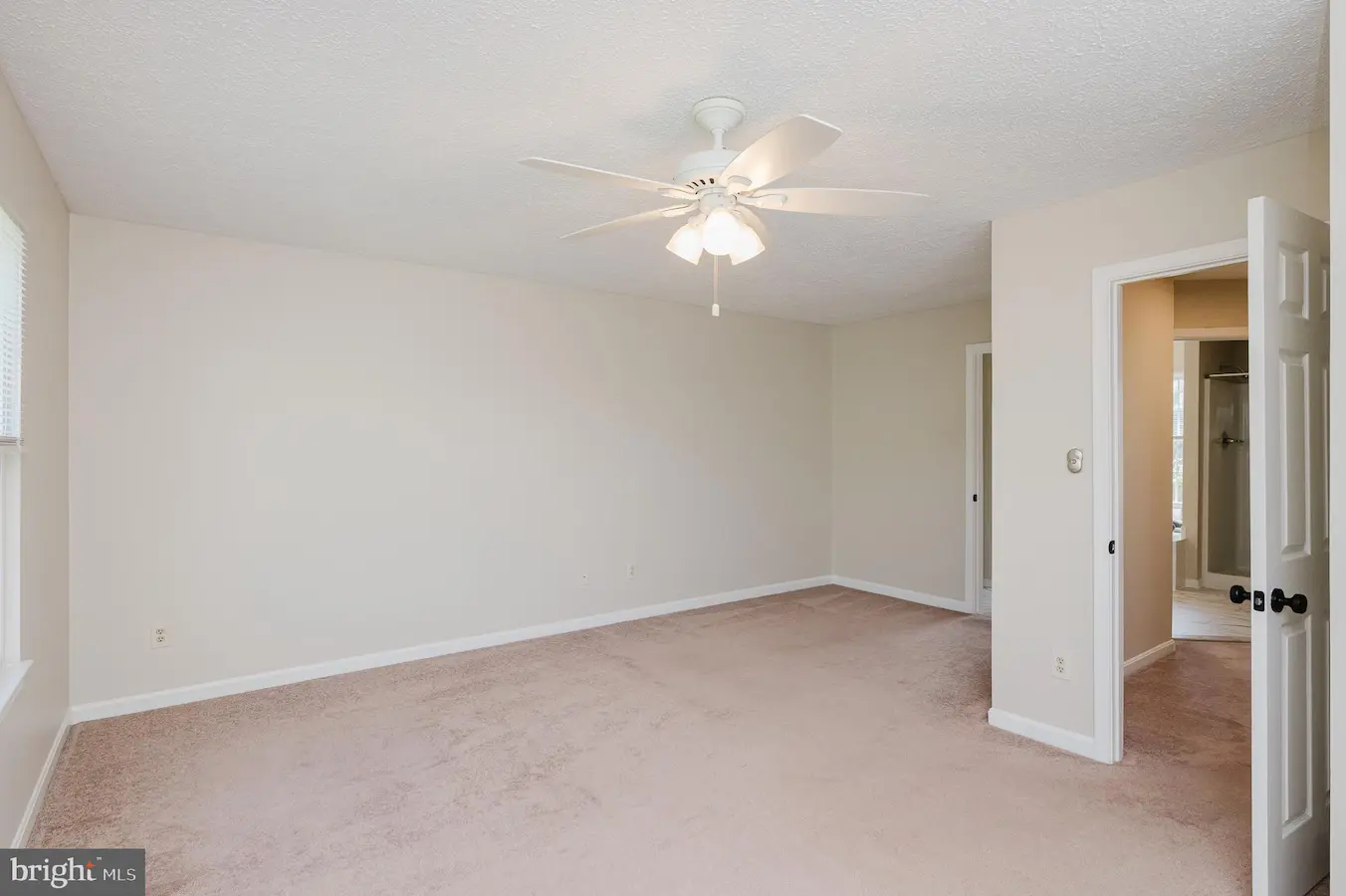 Empty beige bedroom with a white ceiling fan and light, beige carpet, and a doorway to an adjacent bathroom on the right.