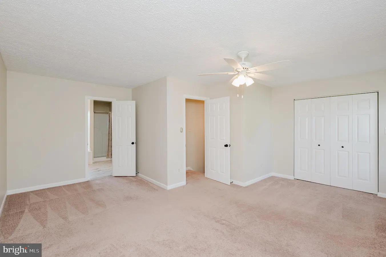 Empty bedroom with beige walls, light carpet, and a white ceiling fan; open doors reveal a bathroom and a hallway, with a double-door closet on the right.