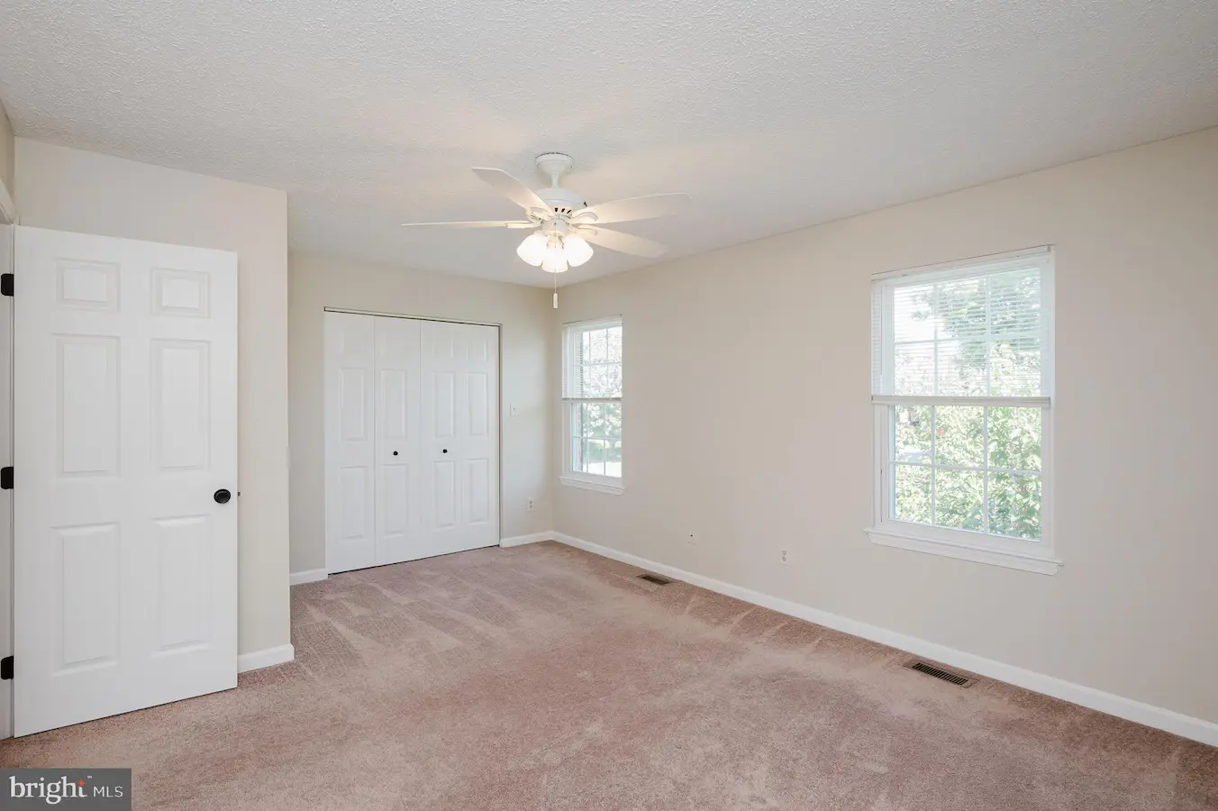 Vacant bedroom with beige walls, pinkish carpet, a white ceiling fan, a double-door closet, and two windows letting in natural light.