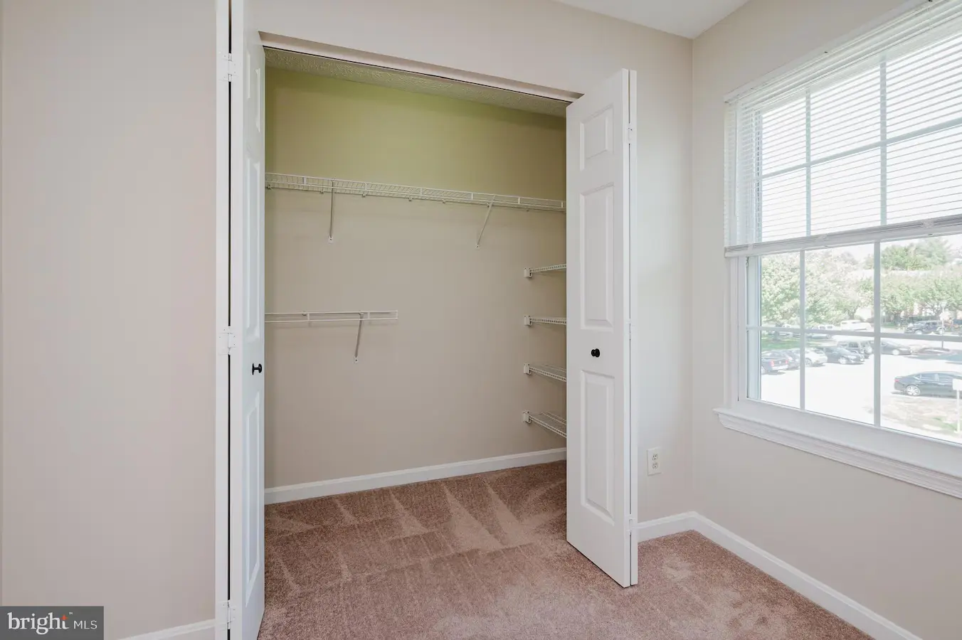 Empty beige closet with white wire shelves and carpeted floor, open double doors beside a window.