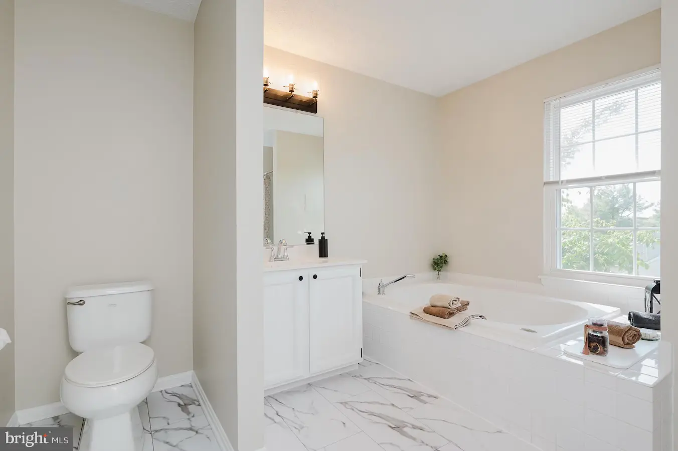Bright modern bathroom with a white vanity, rectangular mirror, and a large soaking tub by a window; tiled marble floor throughout.