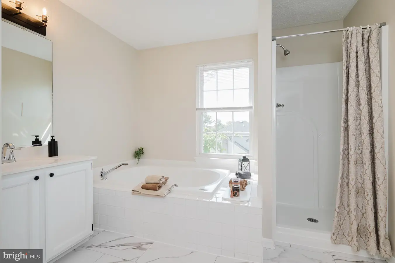 Bright bathroom with a white corner vanity, large bathtub, and a separate shower with a patterned curtain; window lets in natural light.