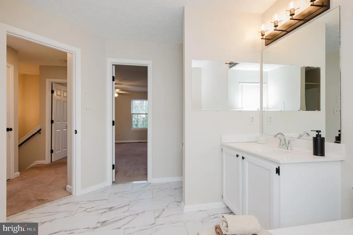 Bright bathroom with white cabinetry and a double sink, large mirrored vanity, and a four-bulb light fixture. Marble floor and beige walls.