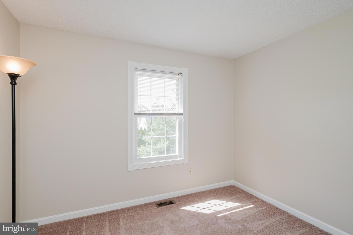 Empty beige room with a white-framed window and blinds; a floor lamp in the left corner.